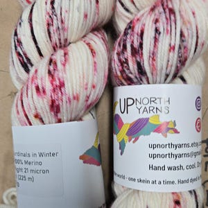 May include: Two skeins of hand-dyed yarn. The yarn is off-white with splashes of red and black. The labels read "Up North Yarns" and include the text "Cardinals in Winter" and "100% Merino".