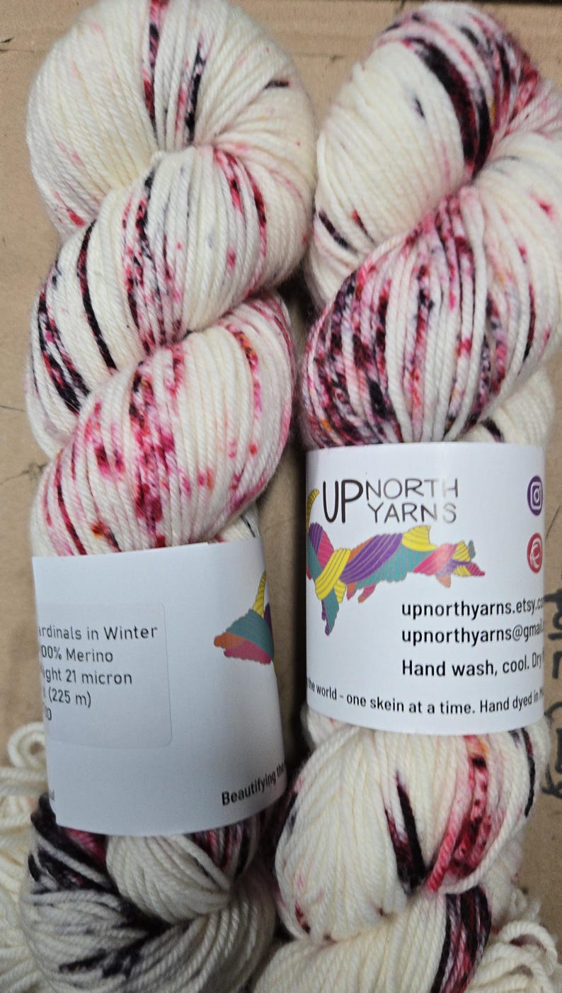 May include: Two skeins of hand-dyed yarn. The yarn is off-white with splashes of red and black. The labels read "Up North Yarns" and include the text "Cardinals in Winter" and "100% Merino".