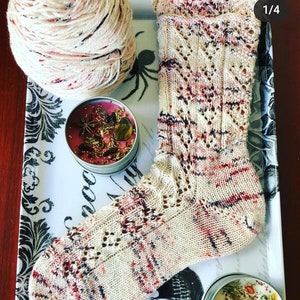 May include: A hand-knitted sock with a cream-colored base and red, black, and purple accents. The sock features a decorative openwork pattern. A ball of yarn and two small candles are also visible on a decorative tray.