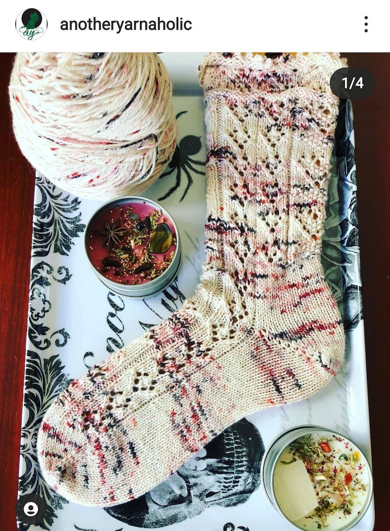 May include: A hand-knitted sock with a cream-colored base and red, black, and purple accents. The sock features a decorative openwork pattern. A ball of yarn and two small candles are also visible on a decorative tray.