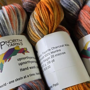 Puede incluir: Varias madejas de lana 100% merino de Up North Yarns en una combinaci&oacute;n de colores variados de carb&oacute;n, naranja y blanco. Cada madeja tiene una etiqueta de papel blanco con el texto Fayette Charcoal Kiln, Superwash 100% Merino, Worsted Weight 20 micron, 100 g 218 yd.