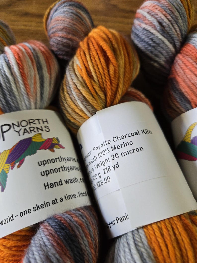 Puede incluir: Varias madejas de lana 100% merino de Up North Yarns en una combinaci&oacute;n de colores variados de carb&oacute;n, naranja y blanco. Cada madeja tiene una etiqueta de papel blanco con el texto Fayette Charcoal Kiln, Superwash 100% Merino, Worsted Weight 20 micron, 100 g 218 yd.