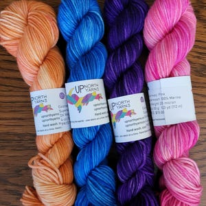 May include: Four skeins of hand-dyed yarn in orange, blue, purple, and pink. Each skein features a white paper label with the text Up North Yarns, social media icons, and product details including 100% Merino wool, 50 g, and 123 yd.
