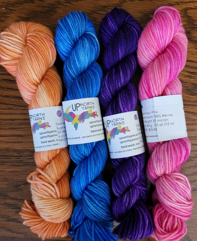 May include: Four skeins of hand-dyed yarn in orange, blue, purple, and pink. Each skein features a white paper label with the text Up North Yarns, social media icons, and product details including 100% Merino wool, 50 g, and 123 yd.