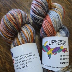 Puede incluir: Dos madejas de hilo Fayette Charcoal Kiln Superwash 100% Merino de peso worsted. El hilo presenta una paleta de colores variados en gris, naranja, amarillo y blanco. Cada madeja tiene una etiqueta de papel blanco con la marca Up North Yarns.