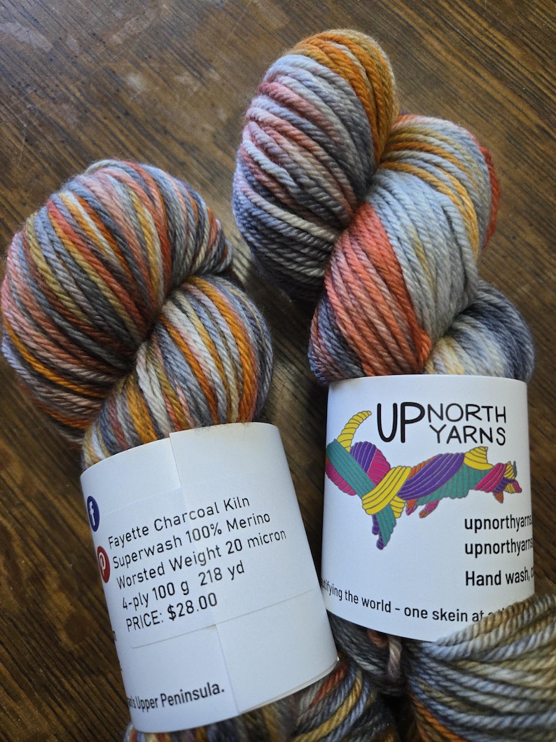 Puede incluir: Dos madejas de hilo Fayette Charcoal Kiln Superwash 100% Merino de peso worsted. El hilo presenta una paleta de colores variados en gris, naranja, amarillo y blanco. Cada madeja tiene una etiqueta de papel blanco con la marca Up North Yarns.
