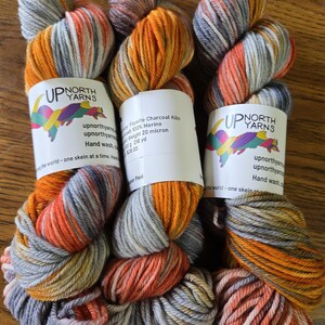 Puede incluir: Tres madejas de hilo te&ntilde;ido a mano con una paleta de colores variados en naranja, gris carb&oacute;n y blanco. Cada madeja tiene una etiqueta de papel blanco con el texto Up North Yarns, Fayette Charcoal Kiln, 100% Merino, 100g y 218 yd.