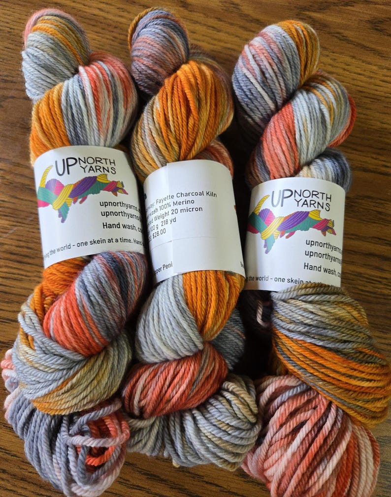Puede incluir: Tres madejas de hilo te&ntilde;ido a mano con una paleta de colores variados en naranja, gris carb&oacute;n y blanco. Cada madeja tiene una etiqueta de papel blanco con el texto Up North Yarns, Fayette Charcoal Kiln, 100% Merino, 100g y 218 yd.