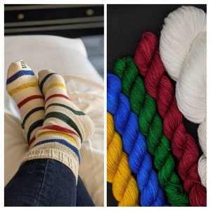 Puede incluir: Un par de calcetines a rayas en blanco, rojo, amarillo y verde. Los calcetines se llevan puestos en las piernas de una persona. La imagen tambi&eacute;n muestra cinco ovillos de lana en amarillo, azul, verde y rojo.