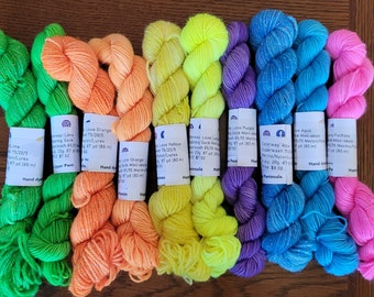 May include: A collection of ten mini-skeins of hand-dyed yarn arranged in a row, featuring vibrant neon colors including lime green, orange, yellow, purple, aqua blue, and fuchsia. Each skein has a white paper label with printed text.