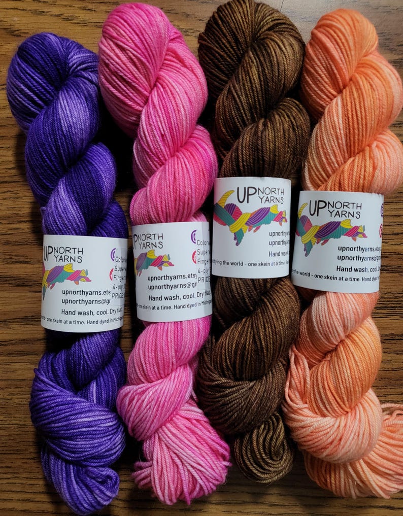 May include: Four skeins of hand-dyed yarn arranged vertically. The colors from left to right are deep purple, bright pink, dark brown, and light peach. Each skein features a white paper label with the text Up North Yarns and care instructions.