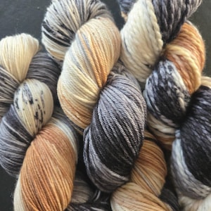 May include: A collection of thick, multi-ply yarn skeins featuring a variegated color palette of cream, charcoal gray, slate, and warm tan. The fibers are twisted into soft, bulky hanks with visible color transitions and speckled patterns.