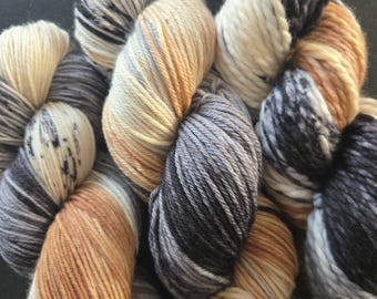 May include: A collection of thick, multi-ply yarn skeins featuring a variegated color palette of cream, charcoal gray, slate, and warm tan. The fibers are twisted into soft, bulky hanks with visible color transitions and speckled patterns.