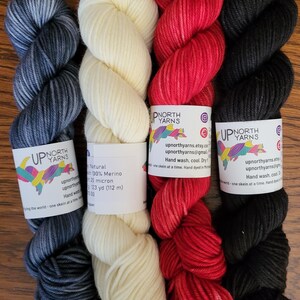 May include: Four skeins of yarn arranged vertically. From left to right: a variegated dark blue and grey skein, a solid cream skein, a variegated red skein, and a solid black skein. Each features a white paper label with the text Up North Yarns.