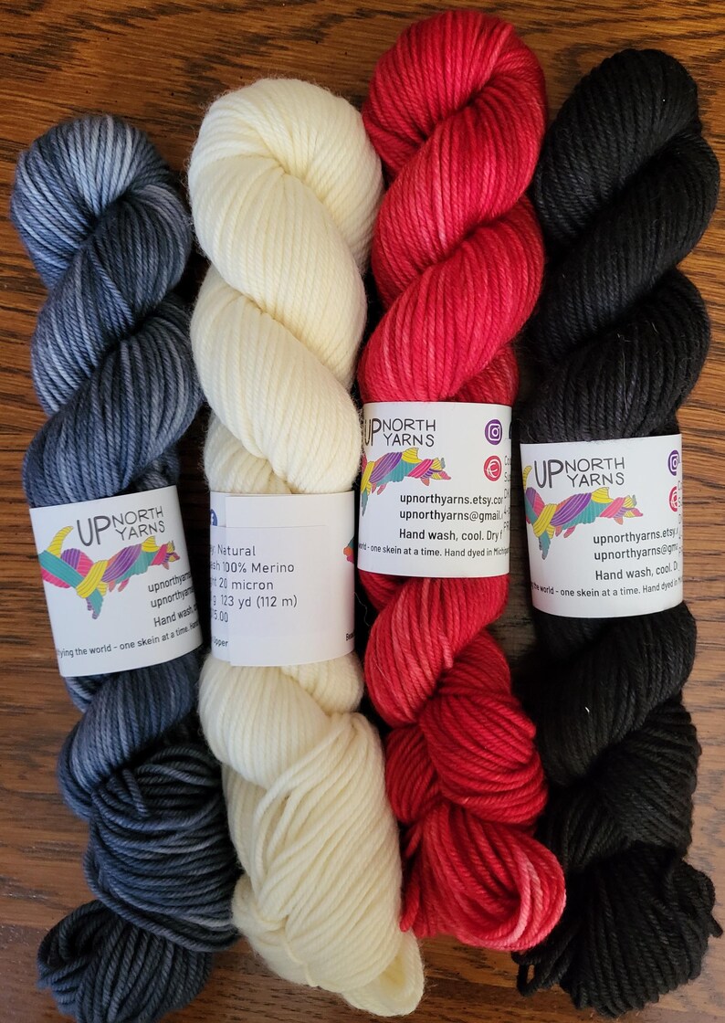 May include: Four skeins of yarn arranged vertically. From left to right: a variegated dark blue and grey skein, a solid cream skein, a variegated red skein, and a solid black skein. Each features a white paper label with the text Up North Yarns.