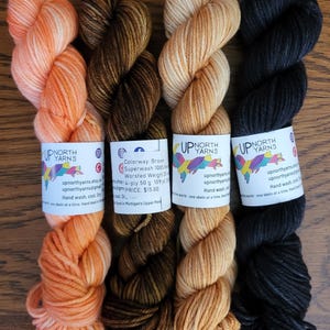 May include: Four skeins of Up North Yarns hand-dyed yarn in orange, dark brown, light tan, and black. Each skein features a white paper label with the brand logo, social media icons, and care instructions. The brown yarn label specifies 50 g and 109 yd.