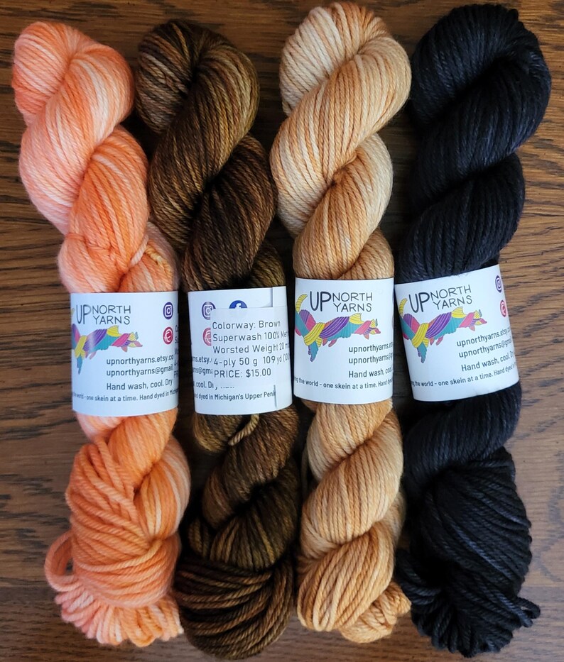 May include: Four skeins of Up North Yarns hand-dyed yarn in orange, dark brown, light tan, and black. Each skein features a white paper label with the brand logo, social media icons, and care instructions. The brown yarn label specifies 50 g and 109 yd.