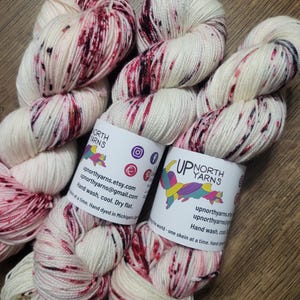 May include: Skeins of hand-dyed yarn in cream with burgundy and black speckles. The yarn is labeled "UP NORTH YARNS" with social media icons and contact information. The label also includes care instructions: Hand wash, cool, dry flat.