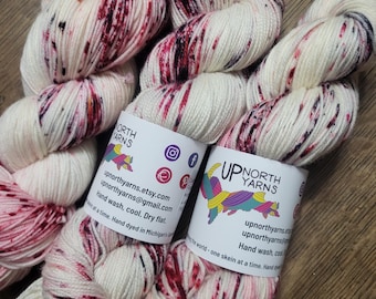 May include: Skeins of hand-dyed yarn in cream with burgundy and black speckles. The yarn is labeled "UP NORTH YARNS" with social media icons and contact information. The label also includes care instructions: Hand wash, cool, dry flat.