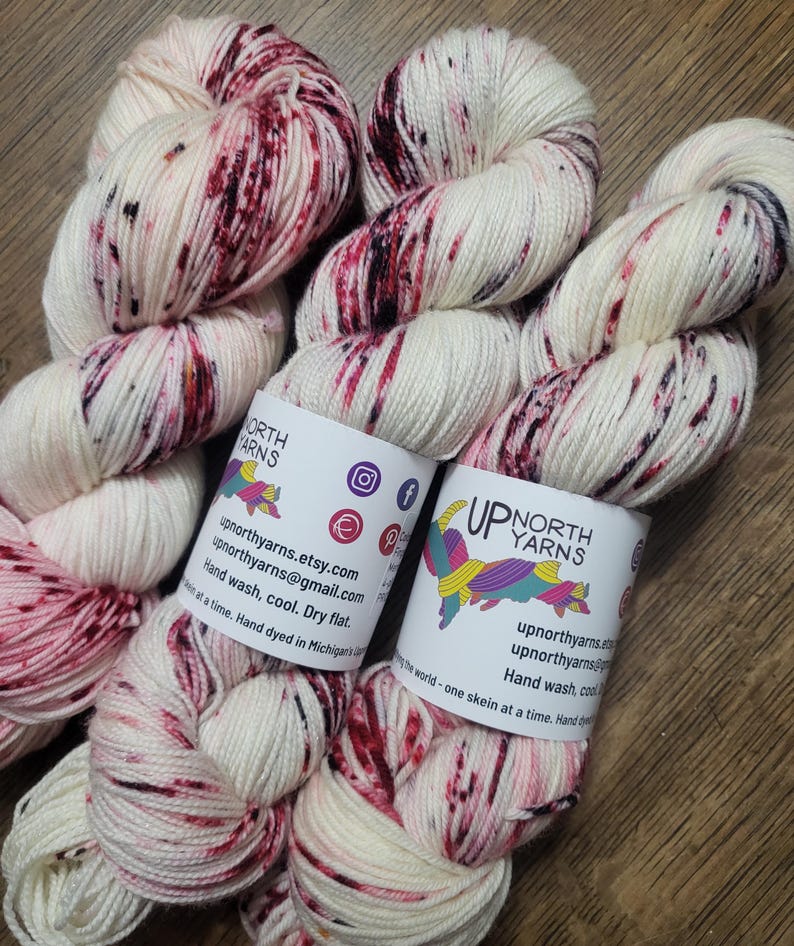 May include: Skeins of hand-dyed yarn in cream with burgundy and black speckles. The yarn is labeled "UP NORTH YARNS" with social media icons and contact information. The label also includes care instructions: Hand wash, cool, dry flat.
