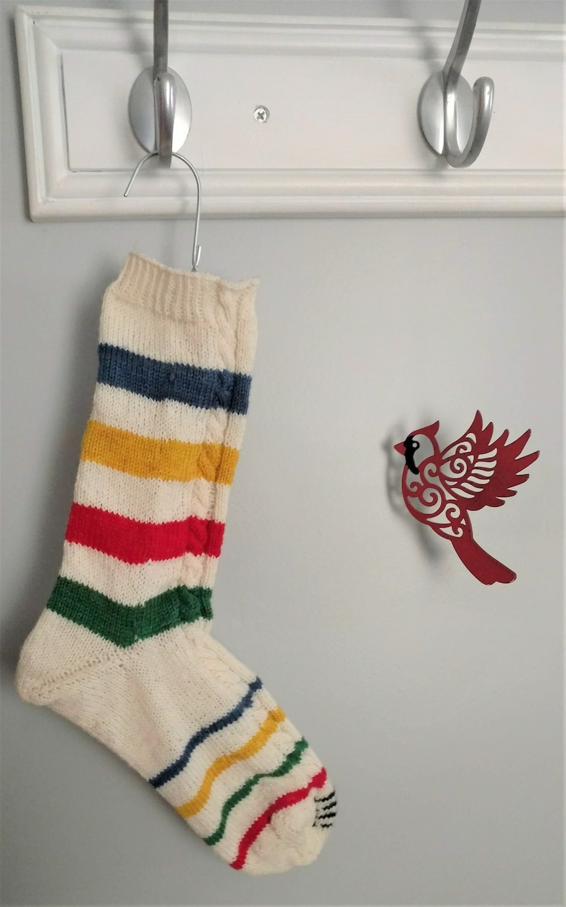 Puede incluir: Un calcet&iacute;n blanco de punto con rayas de colores colgado de un gancho. El calcet&iacute;n tiene rayas azules, amarillas, rojas y verdes. Un recorte de cardenal rojo est&aacute; en la pared detr&aacute;s del calcet&iacute;n.