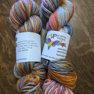 Puede incluir: Dos madejas de hilo te&ntilde;ido a mano de grosor medio en una combinaci&oacute;n de colores gris carb&oacute;n, naranja quemado y amarillo suave. Cada madeja tiene una etiqueta de papel blanco con el texto Up North Yarns, Fayette Charcoal Kiln y 100g.
