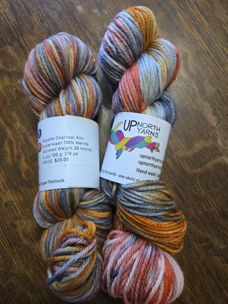 Puede incluir: Dos madejas de hilo te&ntilde;ido a mano de grosor medio en una combinaci&oacute;n de colores gris carb&oacute;n, naranja quemado y amarillo suave. Cada madeja tiene una etiqueta de papel blanco con el texto Up North Yarns, Fayette Charcoal Kiln y 100g.