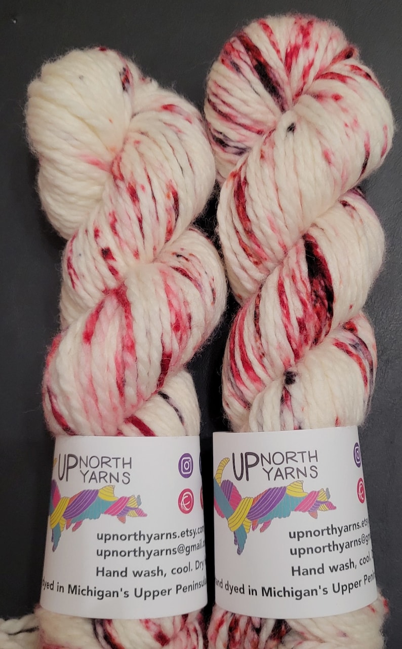 May include: Two skeins of chunky yarn in cream with red and black speckles. Each skein has a label with the text "UP NORTH YARNS" and contact information. The yarn is hand-dyed in Michigan's Upper Peninsula.