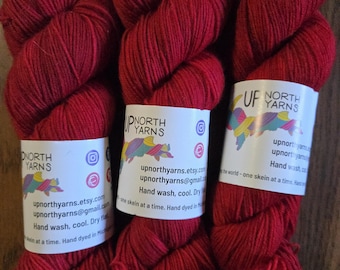 May include: Three skeins of deep red yarn, each wrapped in a white paper label featuring the text Up North Yarns, social media icons, email address, and care instructions stating hand wash, cool, and dry flat, hand dyed in Michigan.
