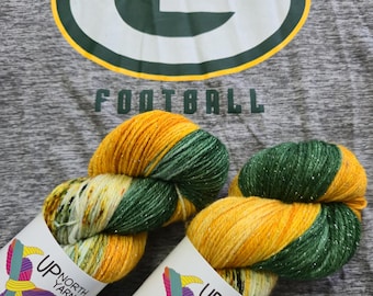 May include: Two skeins of yarn in green, yellow, and white with gold glitter. The yarn is labeled "Up North Yarns" and has the text "beautifying the world - one skein at a time".