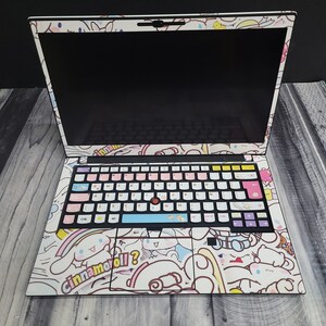 May include: A laptop covered in a white skin featuring a cartoon character design. The keyboard keys are customized with pastel colors. The design includes the text "Cinnamoroll?" on the palm rest area.