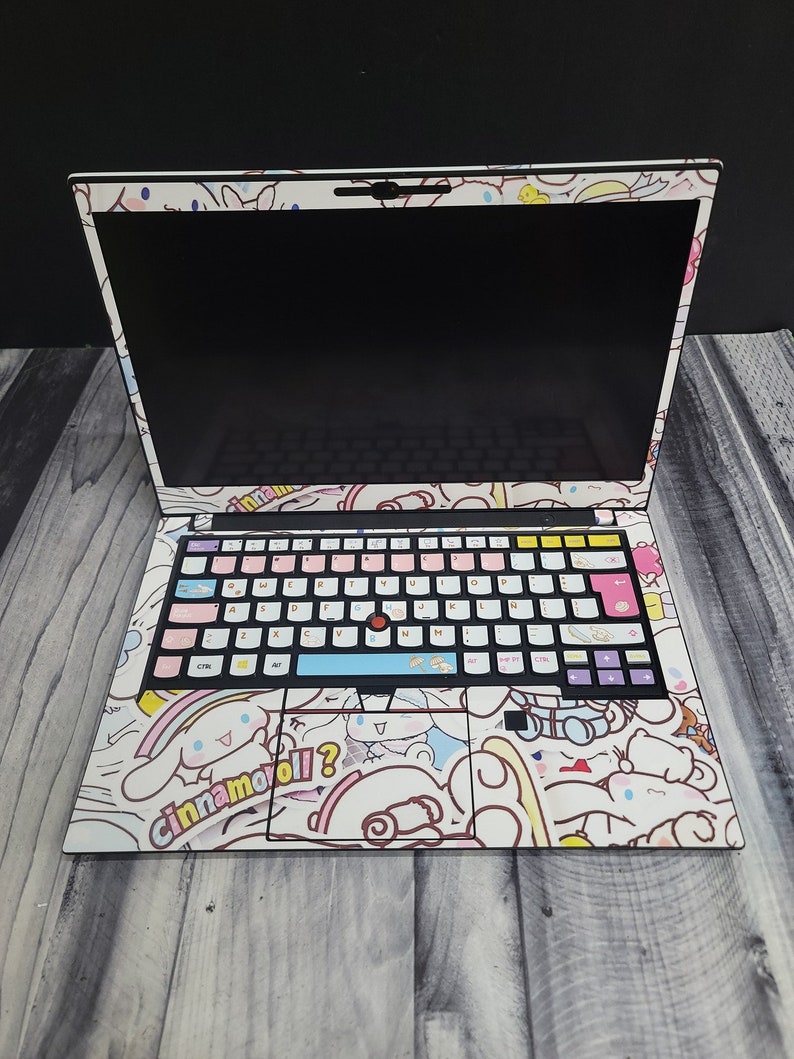 May include: A laptop covered in a white skin featuring a cartoon character design. The keyboard keys are customized with pastel colors. The design includes the text "Cinnamoroll?" on the palm rest area.