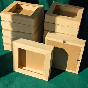 May include: A collection of square wooden shadow box frames with a light natural finish. Each frame features a deep profile, a front-facing opening, and a solid backing panel. One frame is shown from the rear, displaying a metal hanging hook.