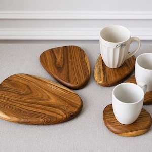 May include: A set of four irregular, pebble-shaped wooden coasters with a natural brown wood grain finish. Three of the coasters are topped with white ceramic mugs, one of which features the word Coffee printed in black script on the side.