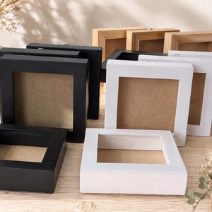 May include: A collection of square shadow box frames in black, white, and natural wood finishes. Each frame features a deep profile with a central opening and a brown backing board, arranged in a group to display their varied colors and depth.