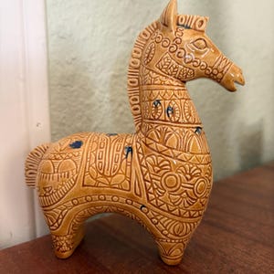 May include: A tan ceramic horse figurine with detailed embossed patterns. The horse features a detailed mane, tail, and body, with blue accents. The figurine is displayed on a wooden surface.