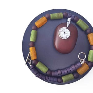 Peut inclure: Un ensemble collier et pendentif fait &agrave; la main. Le collier est compos&eacute; de perles violettes, vertes et orange. Un pendentif ovale marron fonc&eacute; avec un motif en fil d'argent est suspendu au collier. L'ensemble est pr&eacute;sent&eacute; sur une surface circulaire bleu fonc&eacute;.