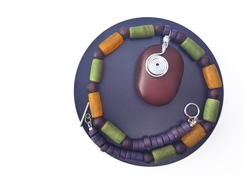 Peut inclure: Un ensemble collier et pendentif fait &agrave; la main. Le collier est compos&eacute; de perles violettes, vertes et orange. Un pendentif ovale marron fonc&eacute; avec un motif en fil d'argent est suspendu au collier. L'ensemble est pr&eacute;sent&eacute; sur une surface circulaire bleu fonc&eacute;.