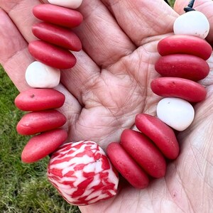 A necklace with red and white beads. The necklace is made up of smooth, oval red beads and round, white beads. A larger, patterned red and white bead is at the centre of the necklace.