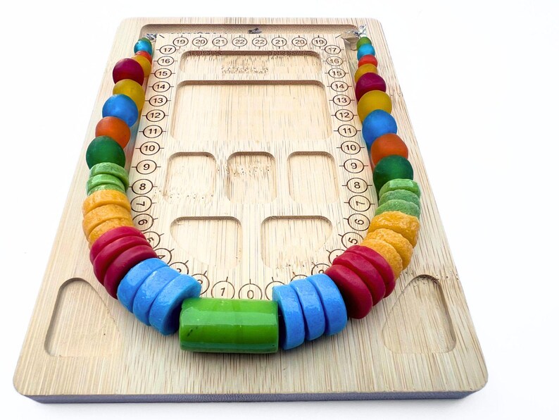 A bamboo bead board with a necklace of colourful recycled glass beads sitting on it. The necklace contains a rainbow of beads in various shapes and sizes, including red, blue, green, orange, and yellow.