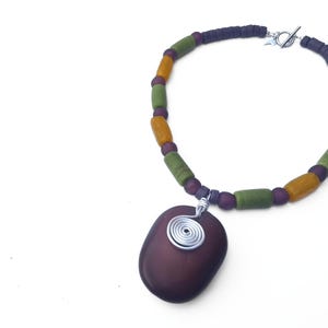 Peut inclure: Collier artisanal avec un pendentif violet fonc&eacute; et une spirale en fil d'argent. Le collier est compos&eacute; de perles vertes, jaunes et violettes. Le fermoir est en argent.