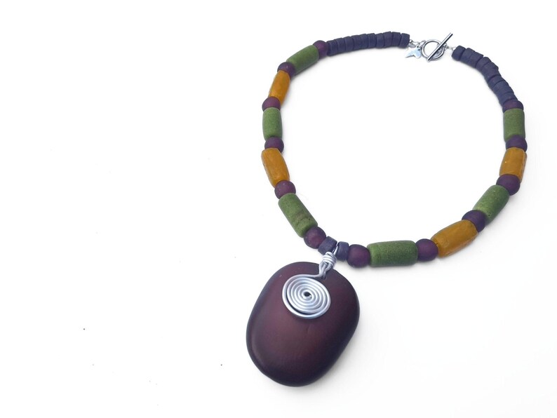 Peut inclure: Collier artisanal avec un pendentif violet fonc&eacute; et une spirale en fil d'argent. Le collier est compos&eacute; de perles vertes, jaunes et violettes. Le fermoir est en argent.
