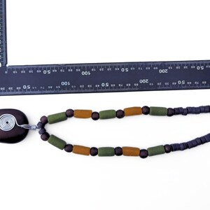 Peut inclure: Collier artisanal avec un pendentif ovale marron fonc&eacute; et une spirale en fil d'argent. Il comporte des perles en bois vert olive et marron, et des perles carr&eacute;es bleu fonc&eacute;. Le collier mesure environ 30 cm de long.