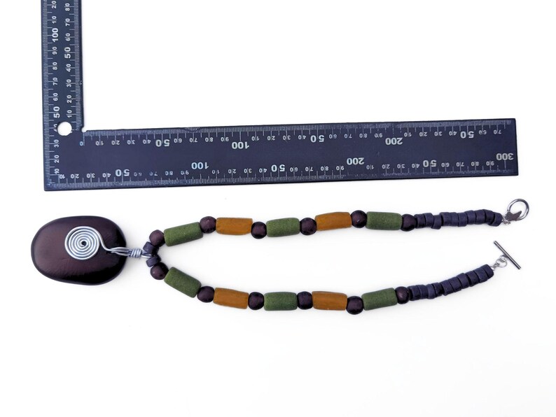 Peut inclure: Collier artisanal avec un pendentif ovale marron fonc&eacute; et une spirale en fil d'argent. Il comporte des perles en bois vert olive et marron, et des perles carr&eacute;es bleu fonc&eacute;. Le collier mesure environ 30 cm de long.