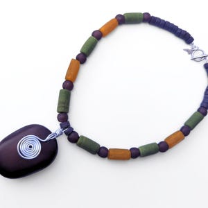 Peut inclure: Un collier artisanal avec un pendentif en pierre violet fonc&eacute; orn&eacute; d'un motif en spirale en fil d'argent. Le collier est compos&eacute; de perles orange, vertes et violet fonc&eacute;, et d'un fermoir argent&eacute;.
