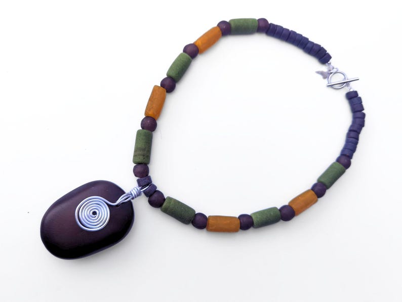 Peut inclure: Un collier artisanal avec un pendentif en pierre violet fonc&eacute; orn&eacute; d'un motif en spirale en fil d'argent. Le collier est compos&eacute; de perles orange, vertes et violet fonc&eacute;, et d'un fermoir argent&eacute;.