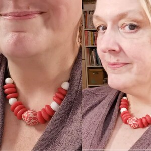 A necklace with red, white, and patterned beads. The necklace is worn around the neck. The beads are a mix of round and disc shapes, creating a bold, colourful statement piece. The necklace is on a black cord.