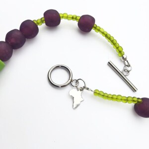 Puede incluir: Un collar con cuentas grandes y redondas de color p&uacute;rpura oscuro intercaladas con cuentas m&aacute;s peque&ntilde;as de color verde claro. El collar tiene un cierre de palanca plateado y un peque&ntilde;o dije con forma de continente africano.