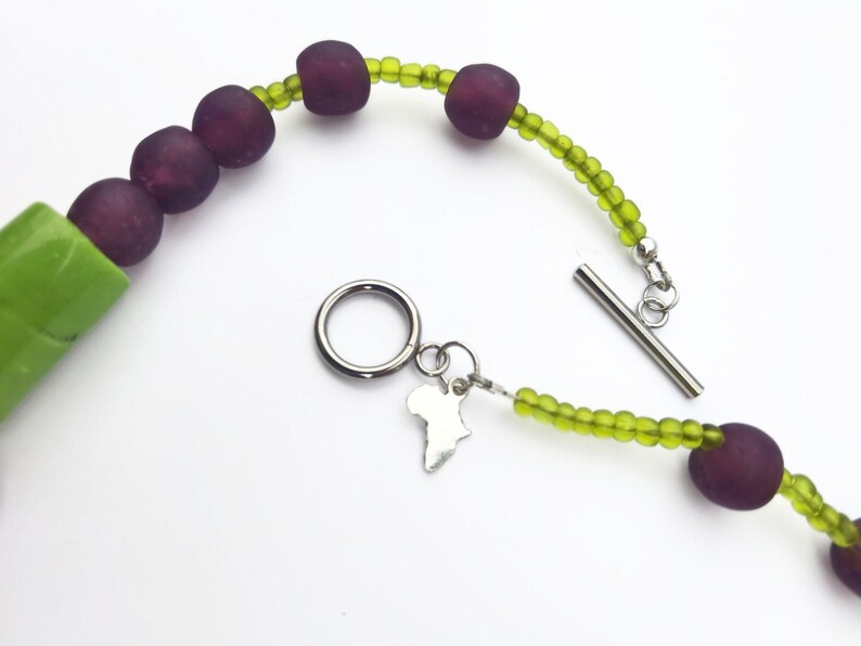 Puede incluir: Un collar con cuentas grandes y redondas de color p&uacute;rpura oscuro intercaladas con cuentas m&aacute;s peque&ntilde;as de color verde claro. El collar tiene un cierre de palanca plateado y un peque&ntilde;o dije con forma de continente africano.