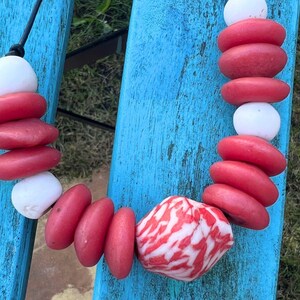 A necklace made with red and white stone beads. The necklace has a black cord and is made up of red, oval-shaped beads and white, round beads. A larger, patterned stone is at the centre.